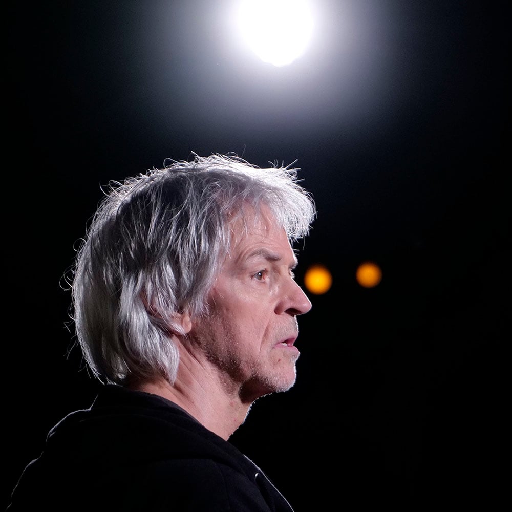 En mann med grått hår og alvorlig ansiktsuttrykk står i profil under et skarpt spotlys, mot en mørk bakgrunn.