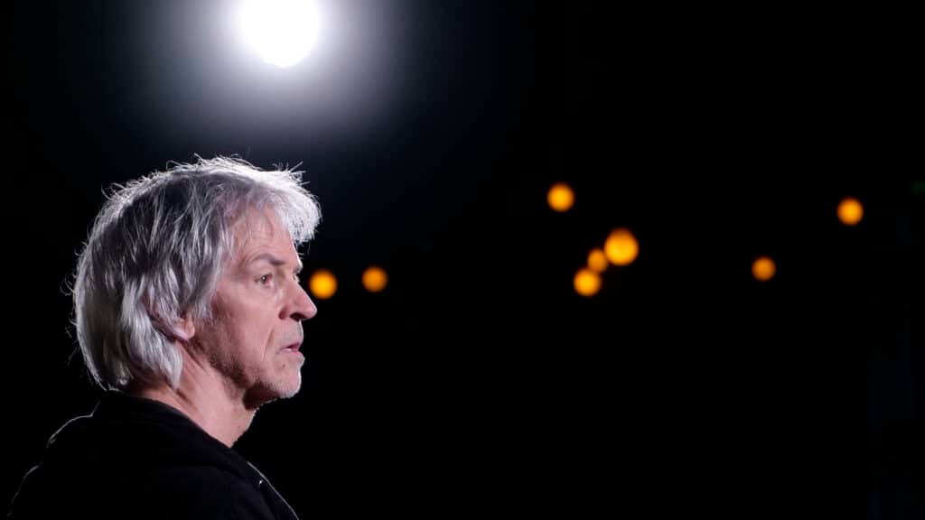 En mann med grått hår står i profil under et skarpt lys, med små oransje lys uskarpt i den mørke bakgrunnen.
