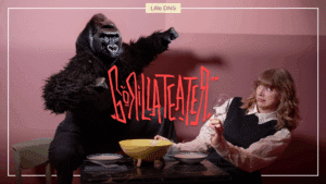 En person i en gorilladrakt sitter ved et bord sammen med en kvinne som holder et glass, med teksten «Lille DNS» og «Gorillateater» på en rosa bakgrunn.