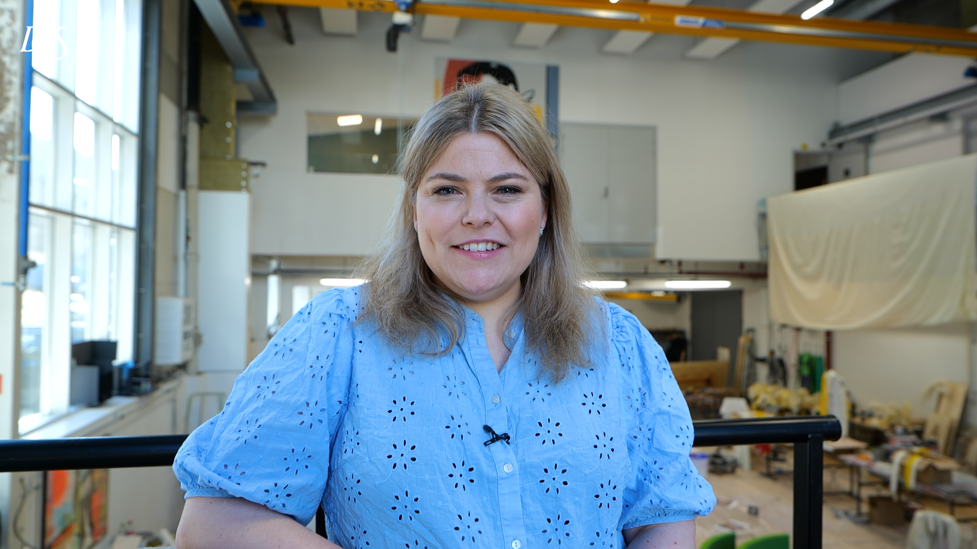 En kvinne med blondt hår og lyseblå bluse står innendørs i et verksted eller industriområde og smiler til kameraet.