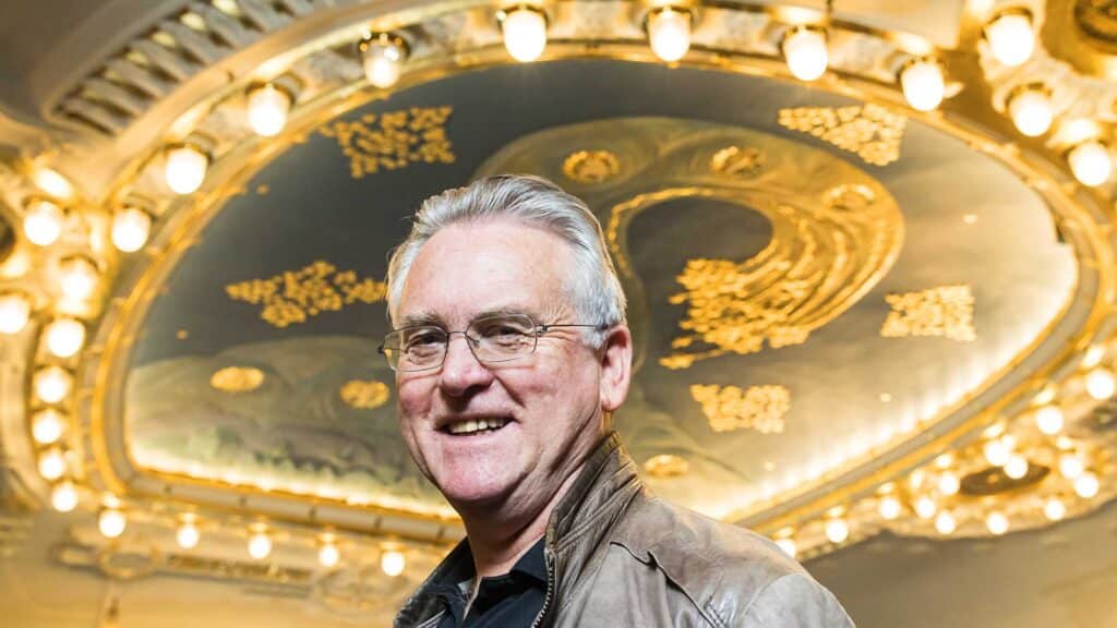 En eldre mann med grått hår og briller i brun jakke står smilende foran et utsmykket, opplyst tak med sirkelformede lamper.