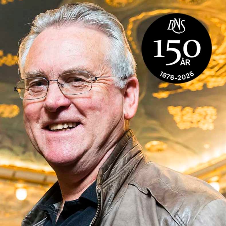 En smilende eldre mann med briller og brun jakke står innendørs. I nærheten står det "150 ÅR 1876-2026" med stiliserte "DNS"-bokstaver over en svart, sirkelformet logo.