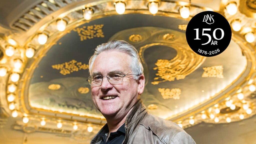 En smilende mann i brun jakke står foran et utsmykket, opplyst tak med et