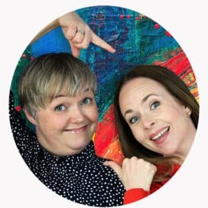 To kvinner smiler og peker på hverandre foran en fargerik, abstrakt bakgrunn.