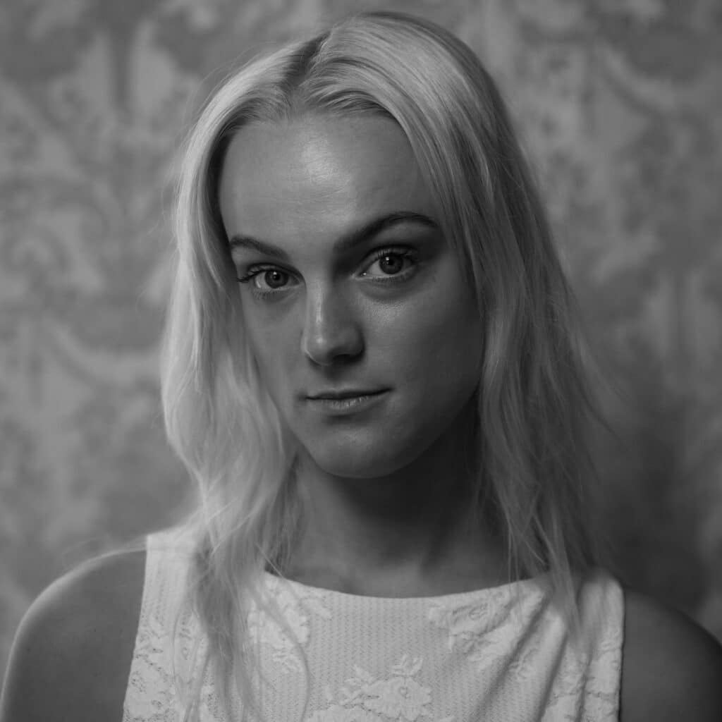 En person med langt, lyst hår og ermeløs, strukturert topp ser inn i kameraet mot en mønstret bakgrunn i svart og hvitt.
