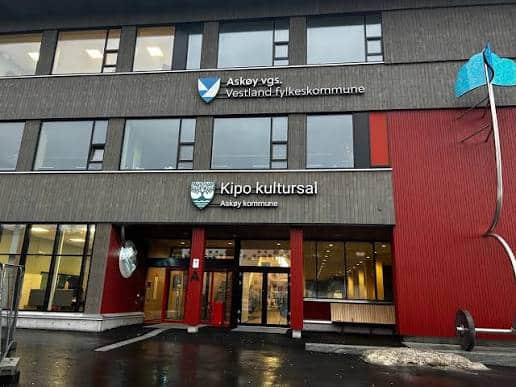 En moderne bygning med skilt for Askøy videregående skole, Vestland fylkeskommune og Kipo kultursal, Askøy kommune, med inngangsdører i glass og vått fortau foran.