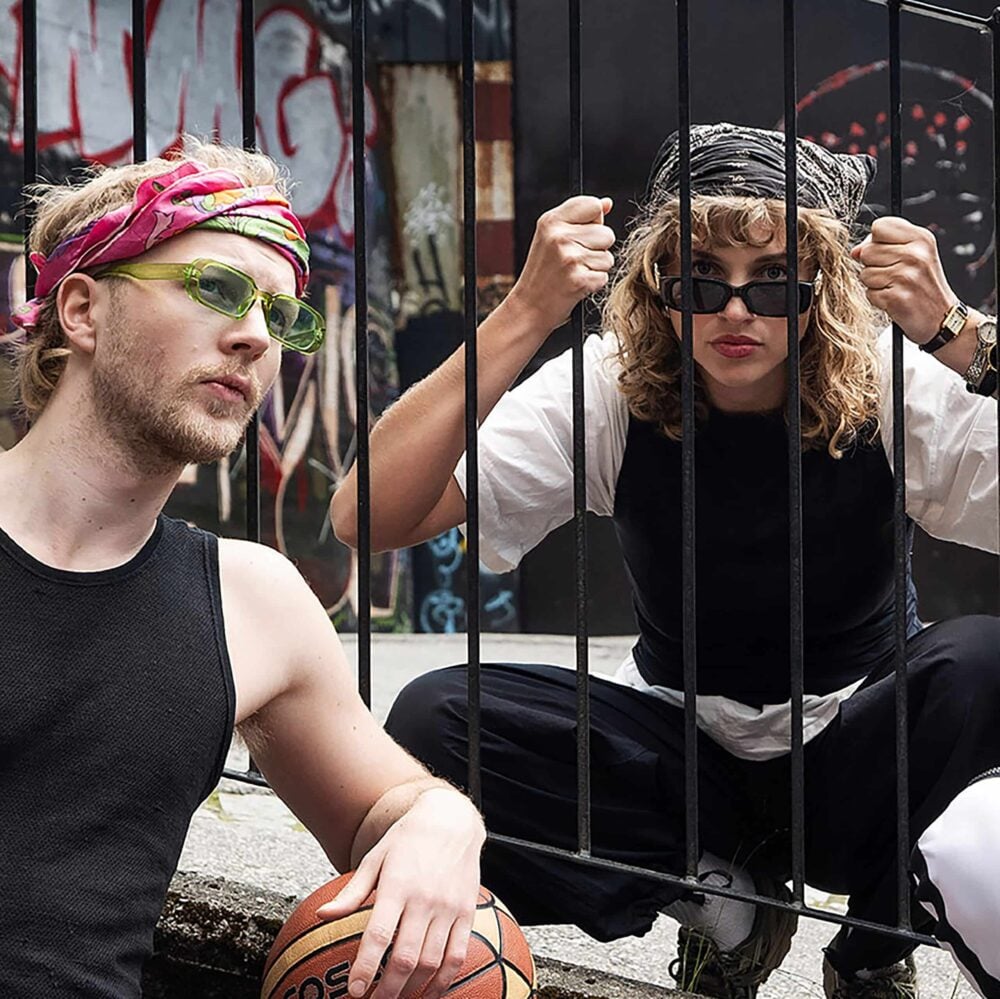 To personer med fargerike pannebånd og solbriller poserer nær et metallgjerde; den ene sitter på huk bak stengene mens den andre sitter og holder en basketball. Urban graffiti er synlig i bakgrunnen.