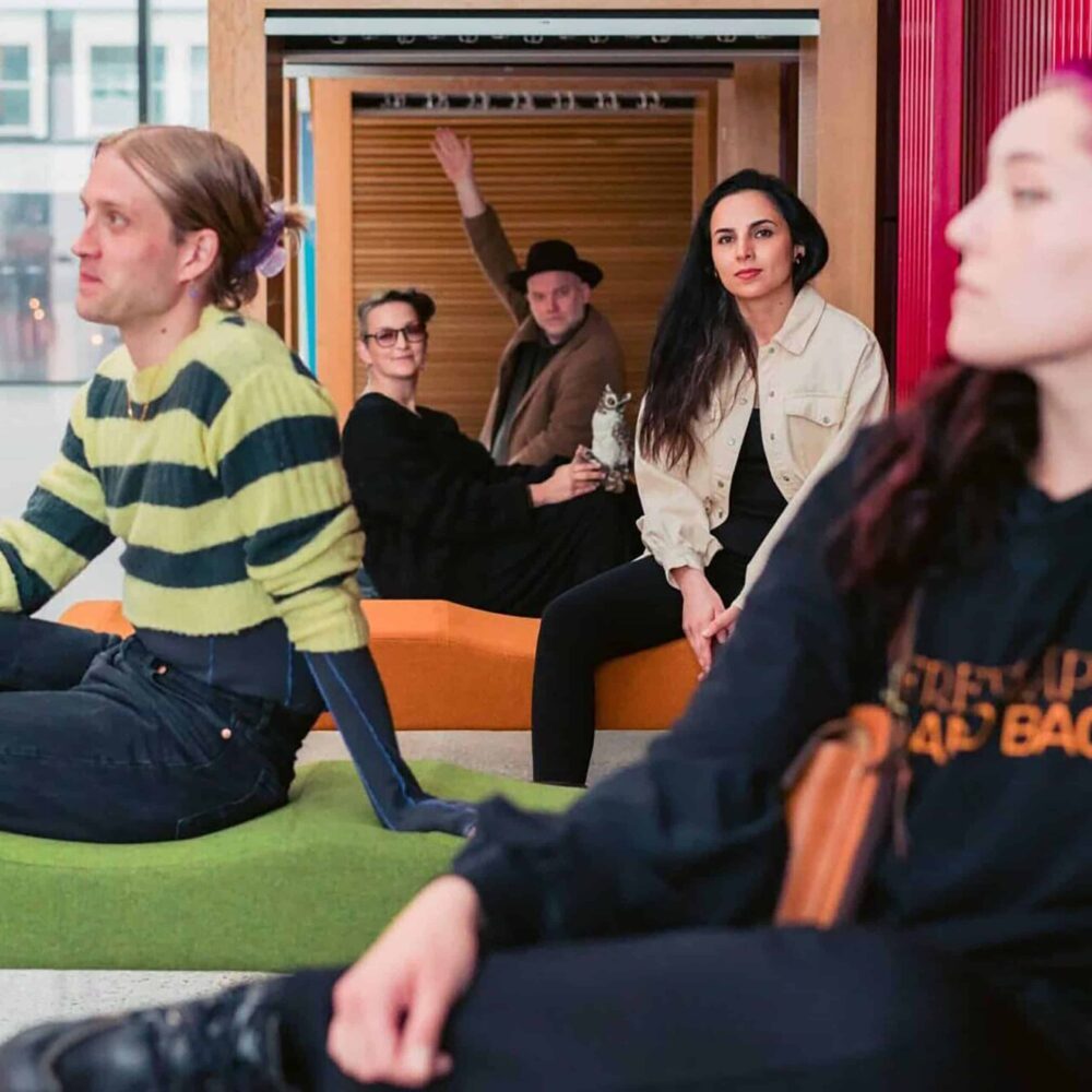 Fem personer sitter eller poserer i et moderne innendørs miljø, noen vender mot kameraet og andre ser bort; én person vinker bakfra.