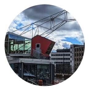 En moderne bygning med en rød, kantete seksjon og store vinduer, metallrammeverk over, og andre kontorbygninger i bakgrunnen under en overskyet himmel.