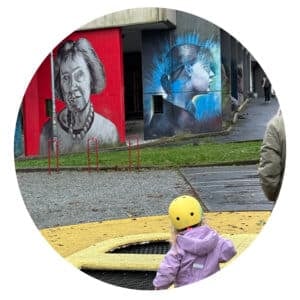 Et barn i gul hjelm og lilla jakke står ved siden av en trampoline, med fargerike veggmalerier av ansikter malt på husvegger i bakgrunnen.