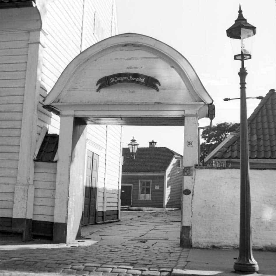 Svart-hvitt-foto av en buet inngang merket "St. Jørgens Hospital", med brosteinsbelagt gate, hvite trebygninger og en gammel gatelykt.