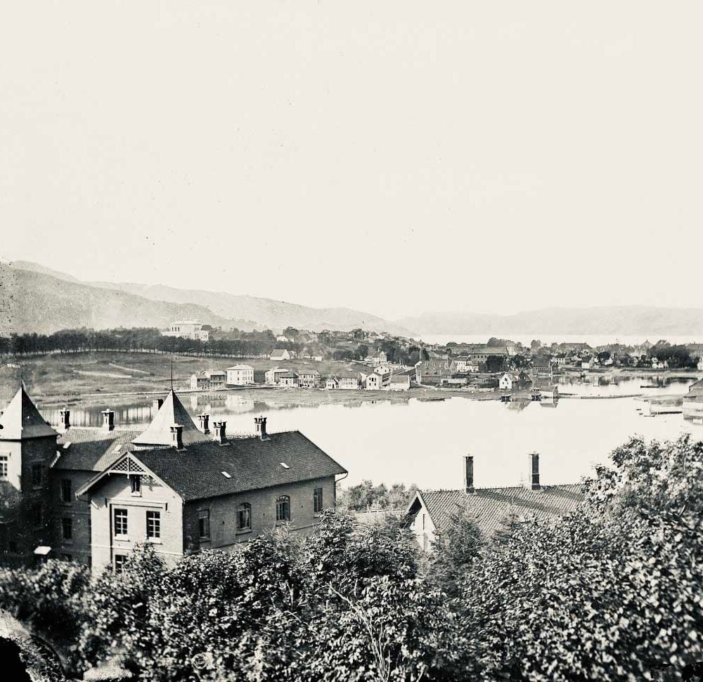 Svart-hvitt-foto av en by ved en innsjø med hus, trær i forgrunnen og fjell i bakgrunnen under en klar himmel.