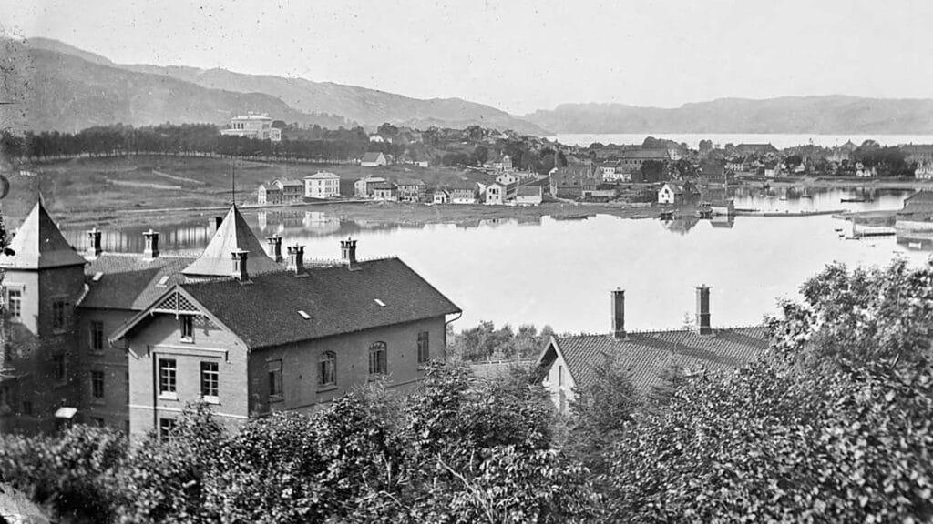 Svart-hvitt-foto av en landsby ved innsjøen med flere hus, trær i forgrunnen og åser i bakgrunnen.