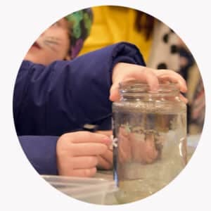 Et barn i blå jakke dypper fingrene i en glasskrukke fylt med vann, mens et annet barn sitter i nærheten.