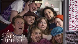 Åtte personer i fargerike kostymer trenger seg inn i en fotoboks og lager overdrevne ansiktsuttrykk. Tekstene «MAESTRO IMPRO» og «FOTOAUTOMAT» vises på bildet.