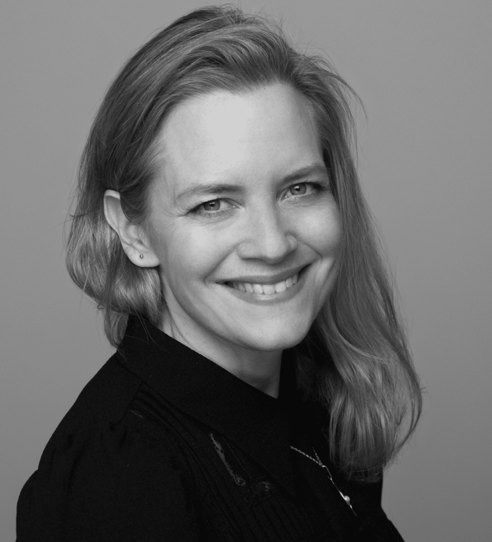 En kvinne med skulderlangt, lyst hår smiler mot kameraet. Hun har på seg en mørk topp og et halskjede, mot en ensfarget bakgrunn.