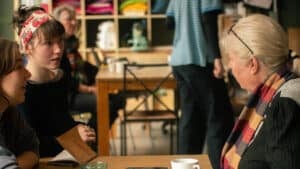 Tre personer sitter ved et bord på en kafé og snakker sammen; én holder en notatbok, og det står krus på bordet. Fargerike hyller og andre gjester er synlige i bakgrunnen.