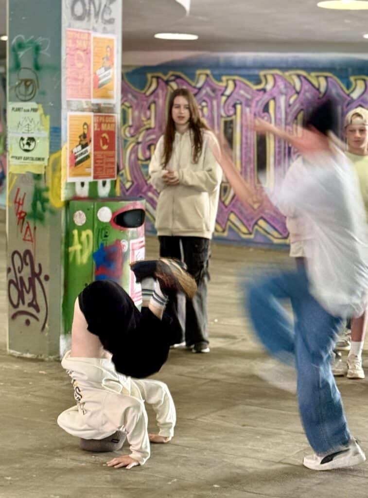 En person står på hodet mens en annen danser i nærheten i et graffitikledd byrom, mens to andre ser på i bakgrunnen.