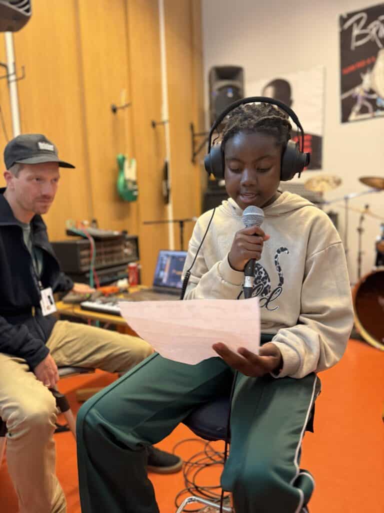En ung person med hodetelefoner holder en mikrofon og leser fra et papir i et musikkstudio, mens en voksen sitter i nærheten. Musikkinstrumenter og utstyr er synlig i bakgrunnen.