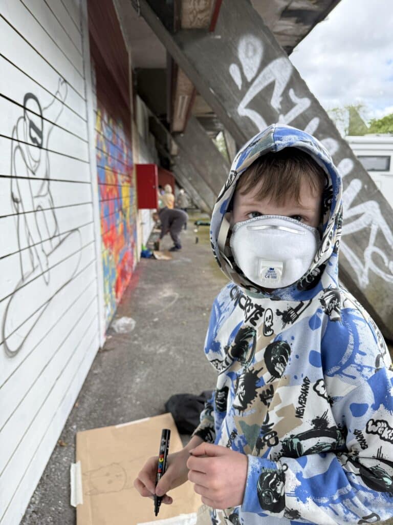 Et barn i hettegenser og ansiktsmaske holder en tusj i nærheten av en vegg med graffiti og en annen person som maler i bakgrunnen.