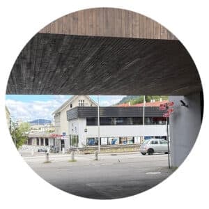 Et gatebilde sett gjennom en sirkelformet ramme viser en bygning, veiskilt, parkerte biler og fjell i bakgrunnen under en delvis overskyet himmel.