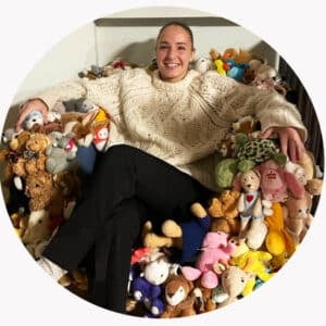 En person iført kremfarget genser sitter på en stol dekket av en stor samling kosedyr og smiler til kameraet.