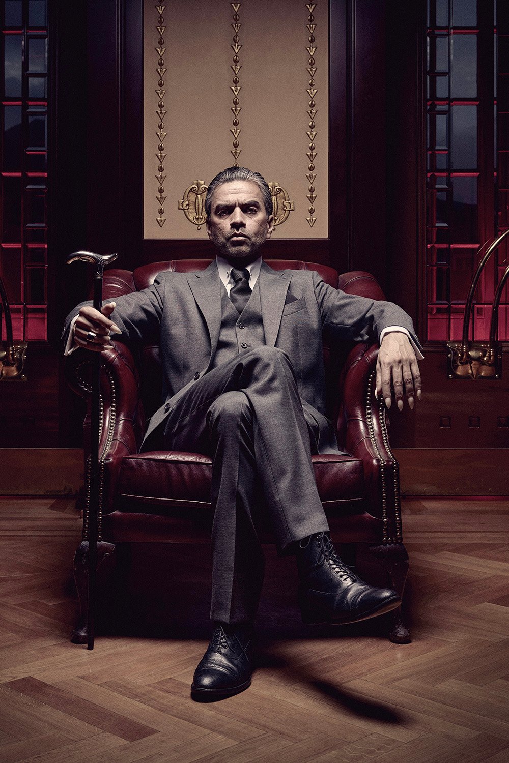 En mann i grå dress sitter selvsikkert i en lenestol i skinn, med stokk og sigar i hånden, med en utsmykket bakgrunn og røde glassmalerier bak seg.
