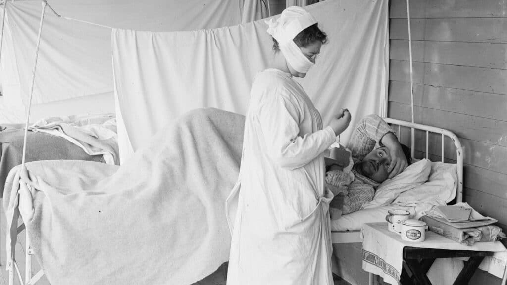 En sykepleier i beskyttelsesklær tar seg av en pasient som ligger i en seng på en provisorisk sykehusavdeling, med medisinsk utstyr på et bord i nærheten.