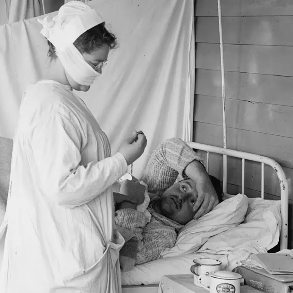 En sykepleier med maske tar seg av en mannlig pasient som ligger i en sykehusseng i et enkelt, sparsomt innredet rom.