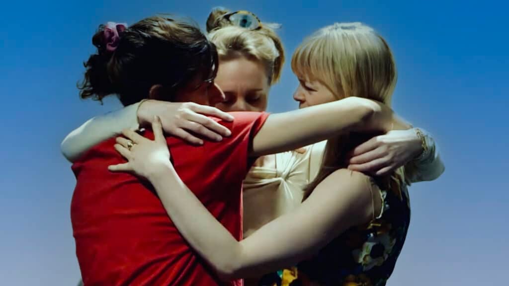 Tre kvinner står tett sammen og omfavner hverandre med armene flettet sammen mot en ensfarget blå bakgrunn.