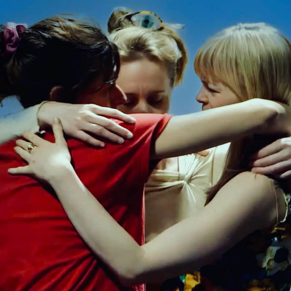 Tre kvinner står tett sammen og omfavner hverandre i en gruppeklem mot en ensfarget blå bakgrunn.