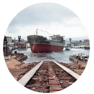 Et stort lasteskip sjøsettes fra et skipsverft, med bygninger og kraner synlige i bakgrunnen under en overskyet himmel.