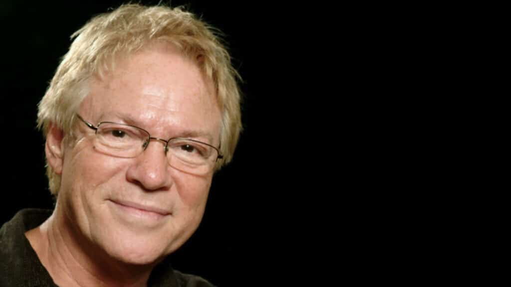 En smilende eldre person med lyst hår og briller er avbildet mot en svart bakgrunn, kledd i en mørk skjorte.