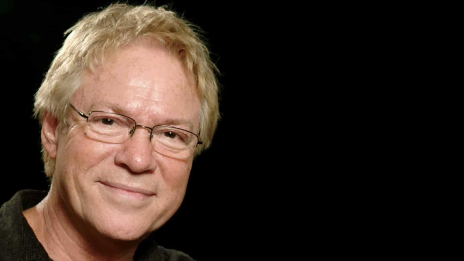 En smilende eldre person med lyst hår og briller er avbildet mot en svart bakgrunn, kledd i en mørk skjorte.