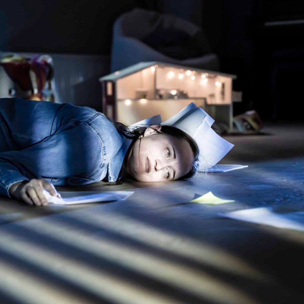 En person ligger på gulvet med papirark rundt seg og på hodet, opplyst av lys fra et dukkehus i bakgrunnen.
