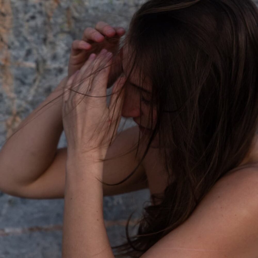 En person med langt, brunt hår holder hendene nær ansiktet, noe som delvis skjuler ansiktstrekkene, mot en grå steinbakgrunn.