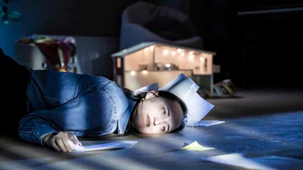 En person ligger på gulvet med papirer spredt rundt seg og på hodet, i et svakt opplyst rom med et dukkehus i bakgrunnen.