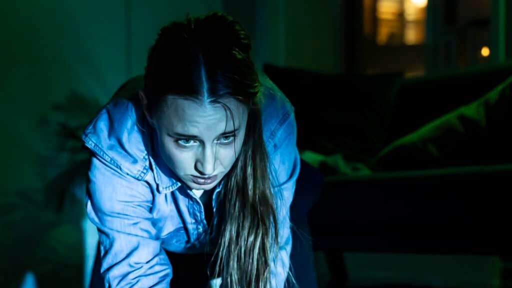 En kvinne med langt hår, iført jeansskjorte, står på alle fire innendørs og ser opp med et alvorlig uttrykk. Belysningen er dunkel og har et blågrønt skjær.