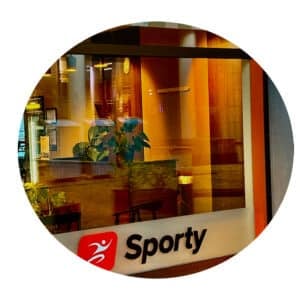 Et glassvindu viser et varmt opplyst interiør med planter og møbler. Under vinduet er det et skilt med et rødt ikon og ordet "Sporty".