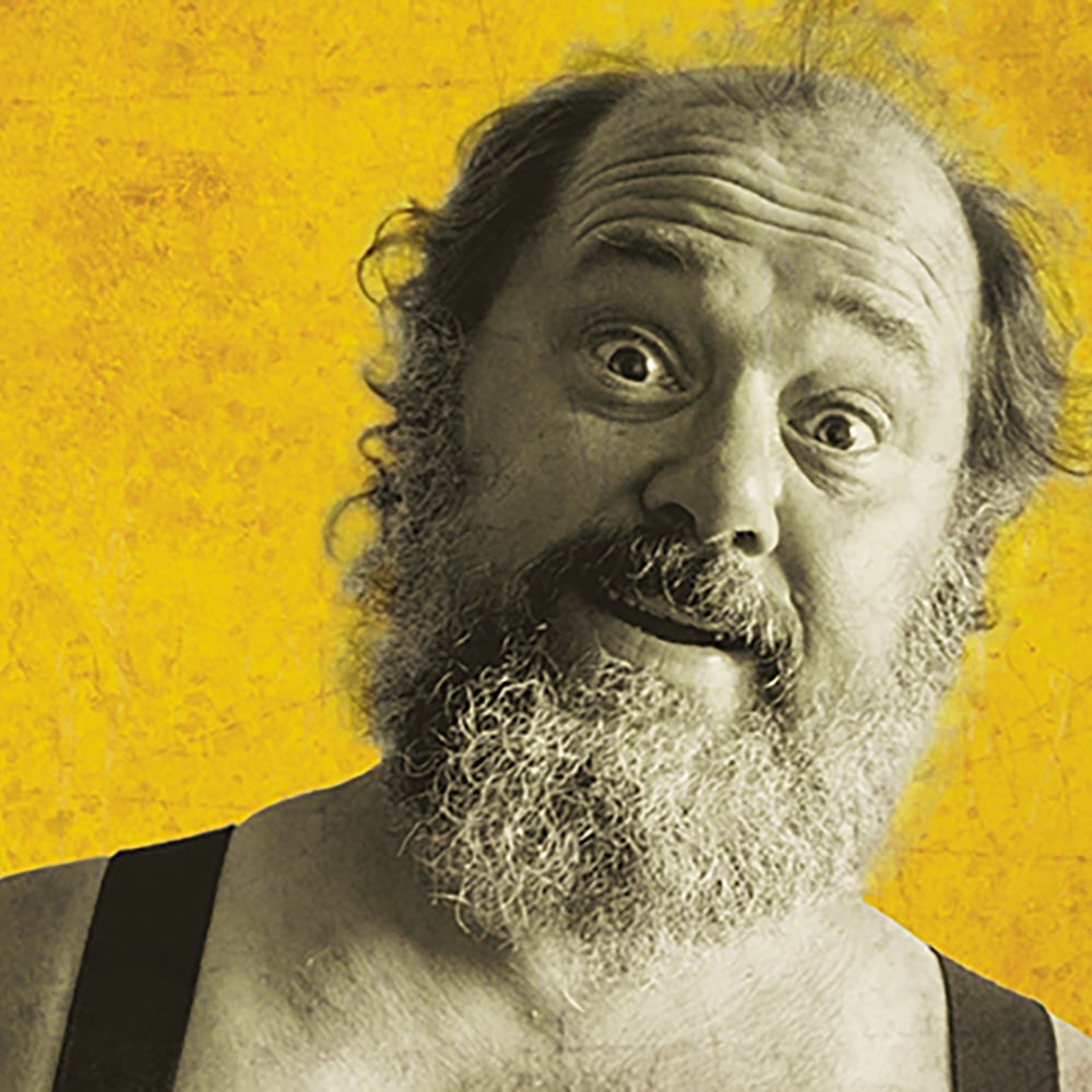 En skjeggete mann med et overrasket ansiktsuttrykk står foran en strukturert gul bakgrunn.