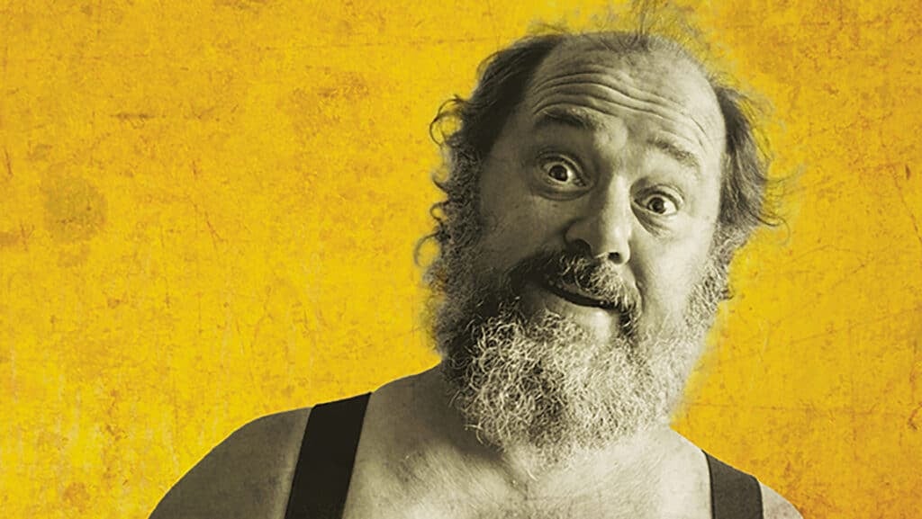 En skjeggete mann med vikende hårfeste og svarte bukseseler ser overrasket inn i kameraet mot en gul bakgrunn.