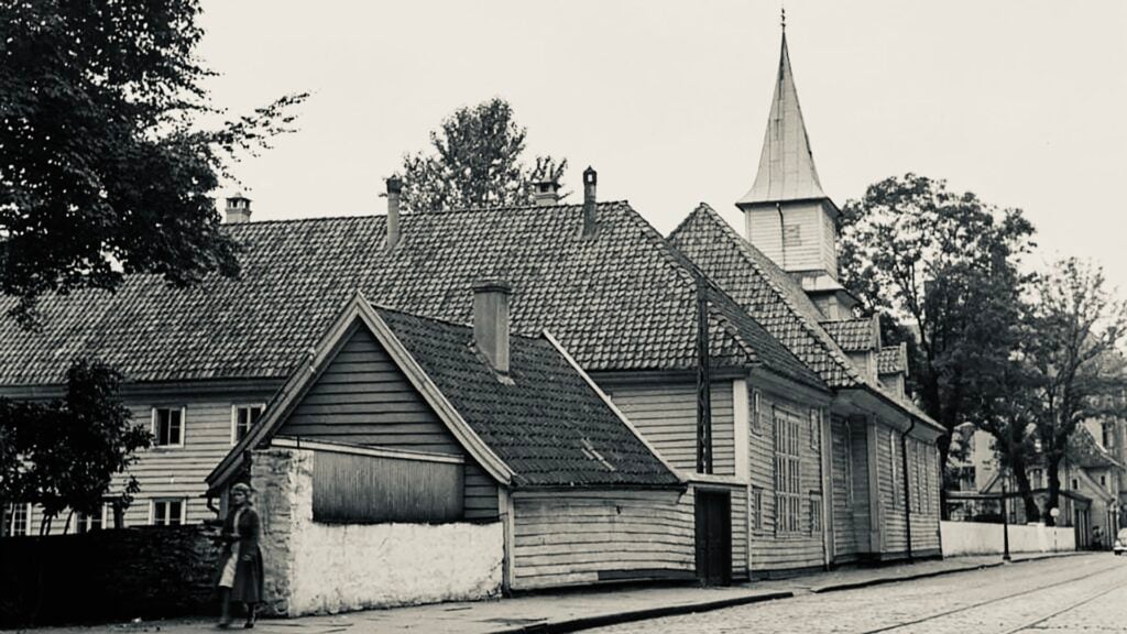 Svart-hvitt-foto av en trekirke med bratt tak og et lite tårn; en person går på fortauet i forgrunnen.