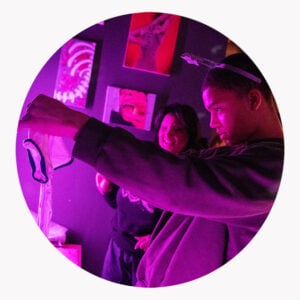 En ung person undersøker et klesplagg i lilla belysning, med en annen person i bakgrunnen og fargerike kunstverk på veggen.