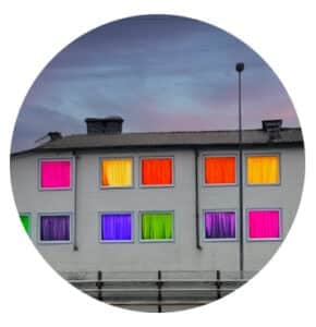 En hvit bygning med flere vinduer, hvert av dem dekket av fargerike gardiner i rosa, oransje, gult, lilla og grønt, under en overskyet himmel.